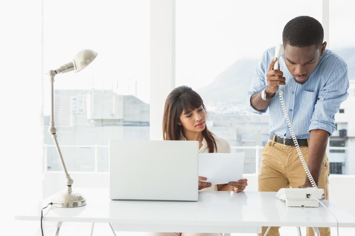 A man in a blue shirt talks on a phone while standing beside a woman in an office. She is seated, holding papers, next to a laptop and a lamp. Bright, professional setting.